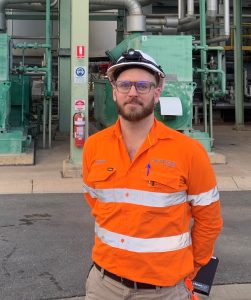 A CleanCo employee named Andrew wears orange high visibility clothing, a hard hat and safety glasses, and stands in front of Swanbank gas-fired power station.