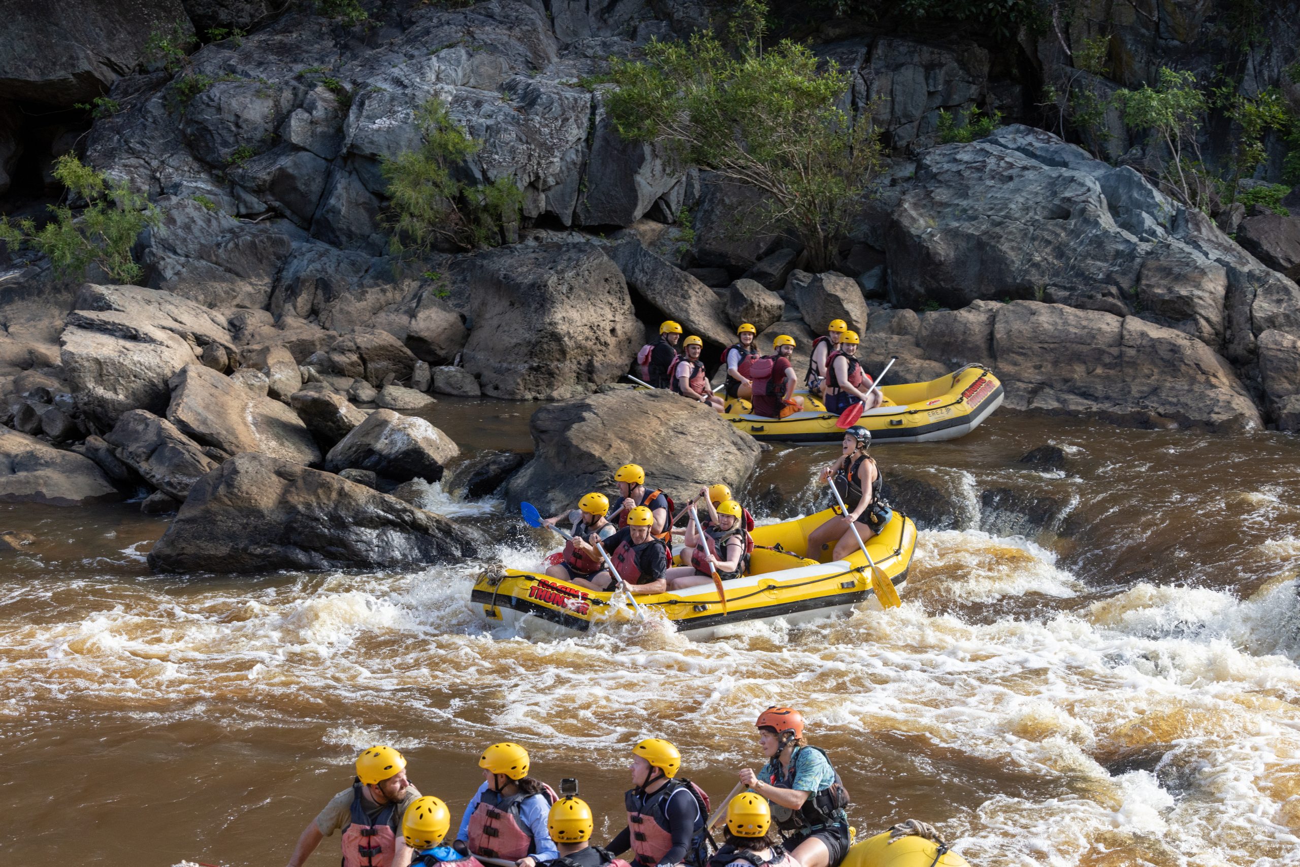 Three groups of people river rafting near the Barron Gorge. There are rapids and they are navigating through the rocks in yellow rafts.