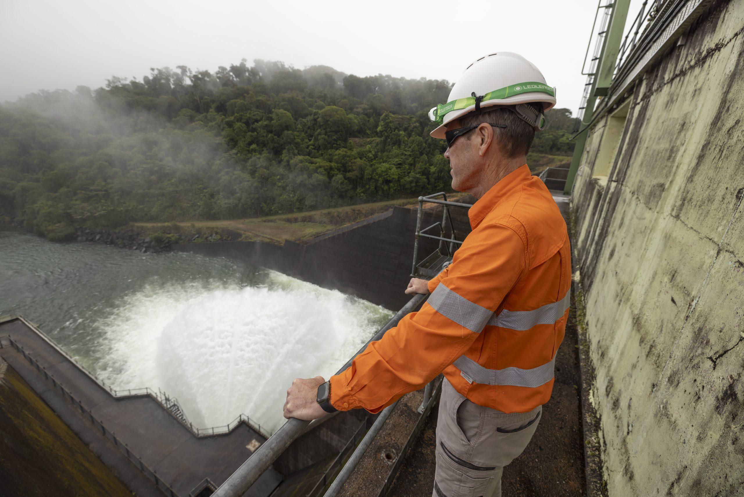 A close-up shot of a CleanCo worker observing the Kareeya and Koombooloomba dam. He is wearing safety gear, has his hands on a rail as he observes the gushing water from the dam wall below.