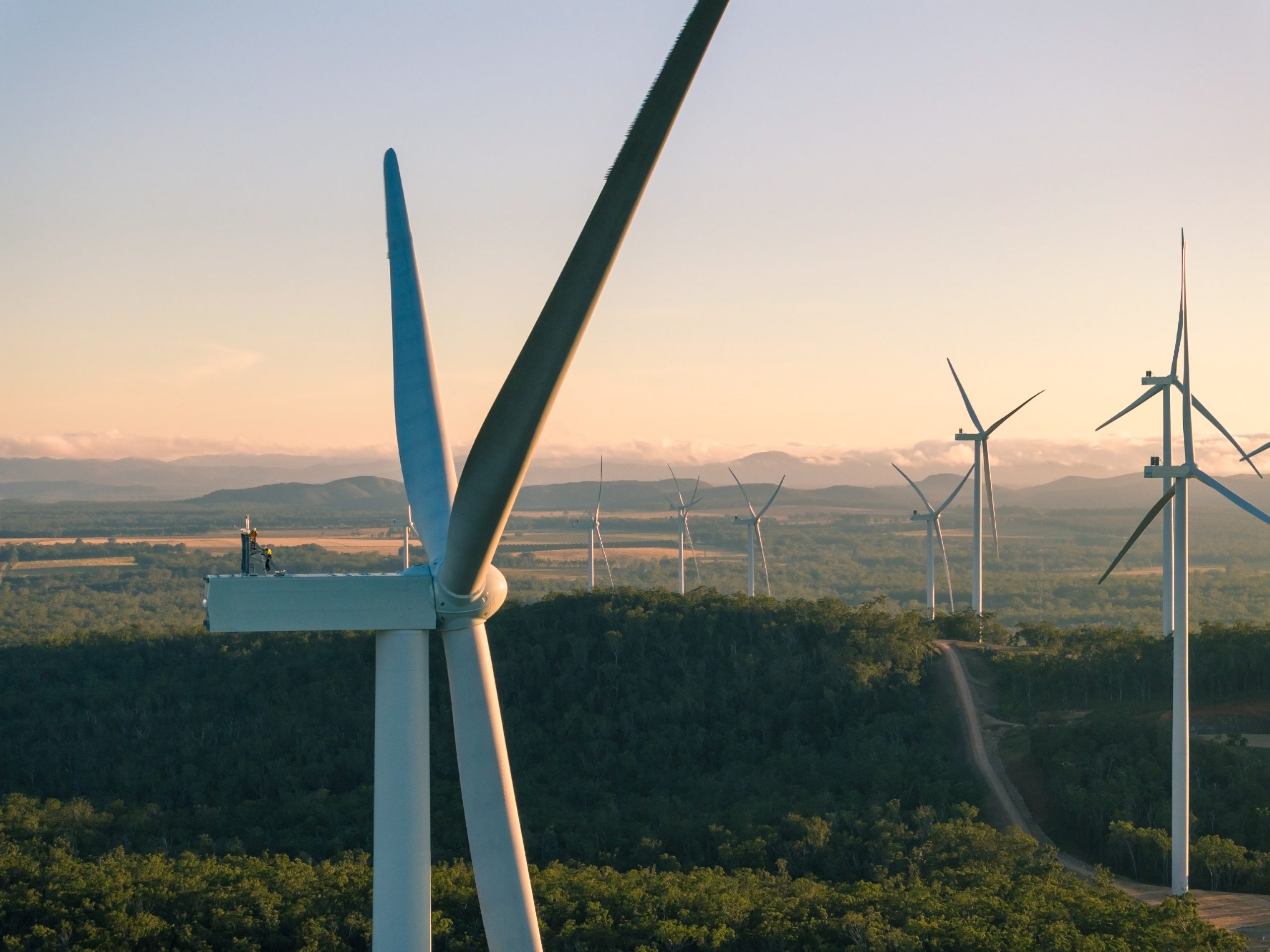 A close-up of a wind turbine at the Kaban Green Power Hub. The close-up is of the propellers and workers that are doing maintenance on the propellers, high above the ground. You can see other wind turbines in the distance.