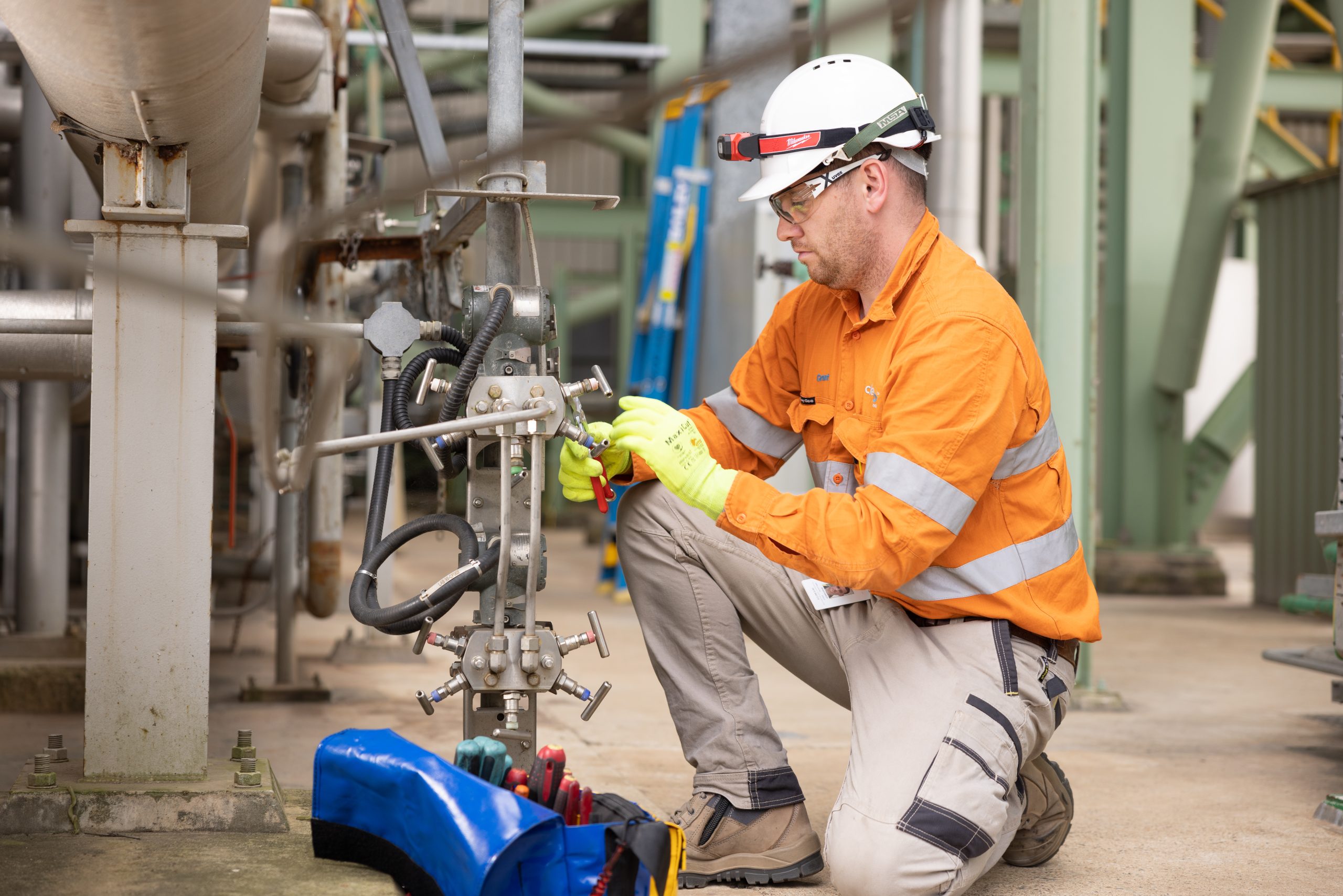 A CleanCo worker performing maintenance on infrastructure in the Swanbank Power Station. He is wearing safety gear, kneeling and holding a wrench.