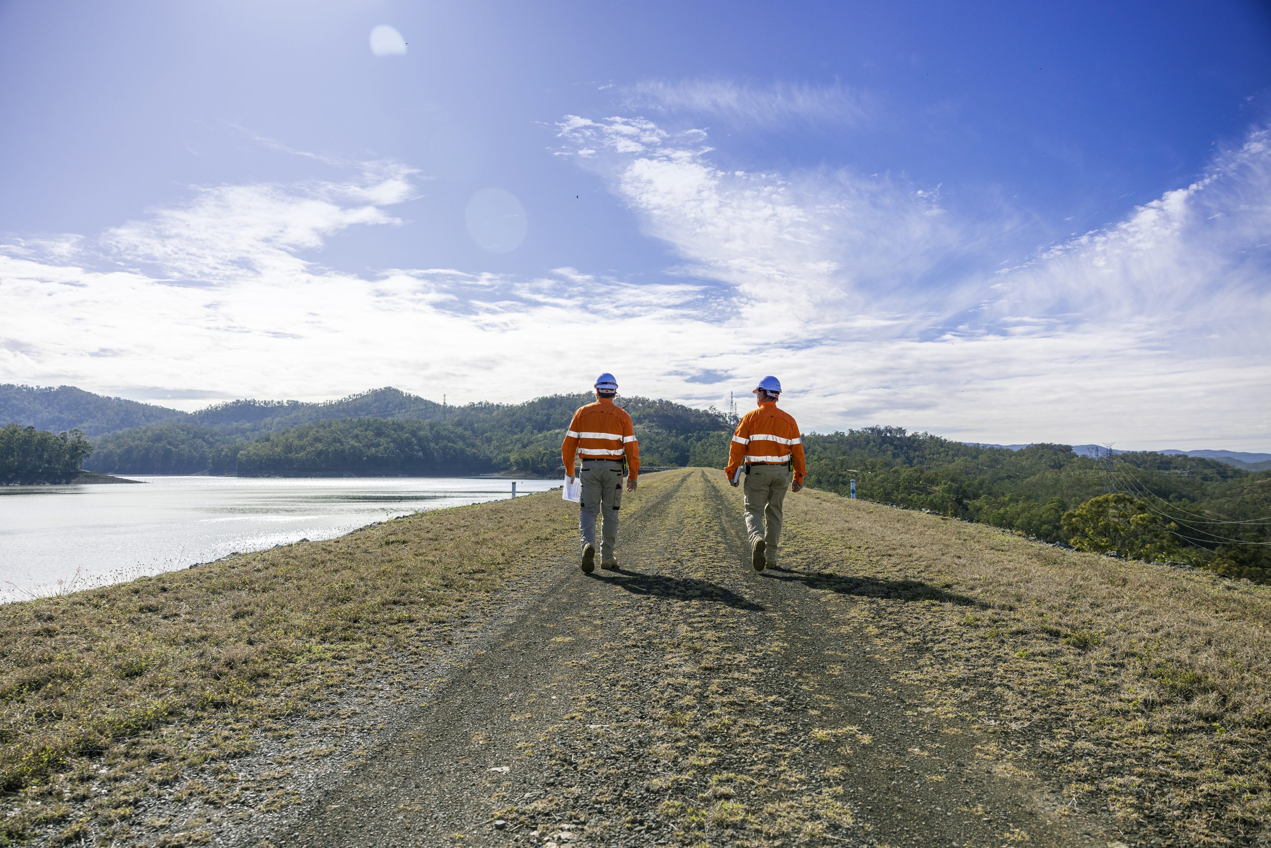 Two CleanCo workers walking along a road next to Wivenhoe Dam. They are wearing safety gear and walking away from the camera.