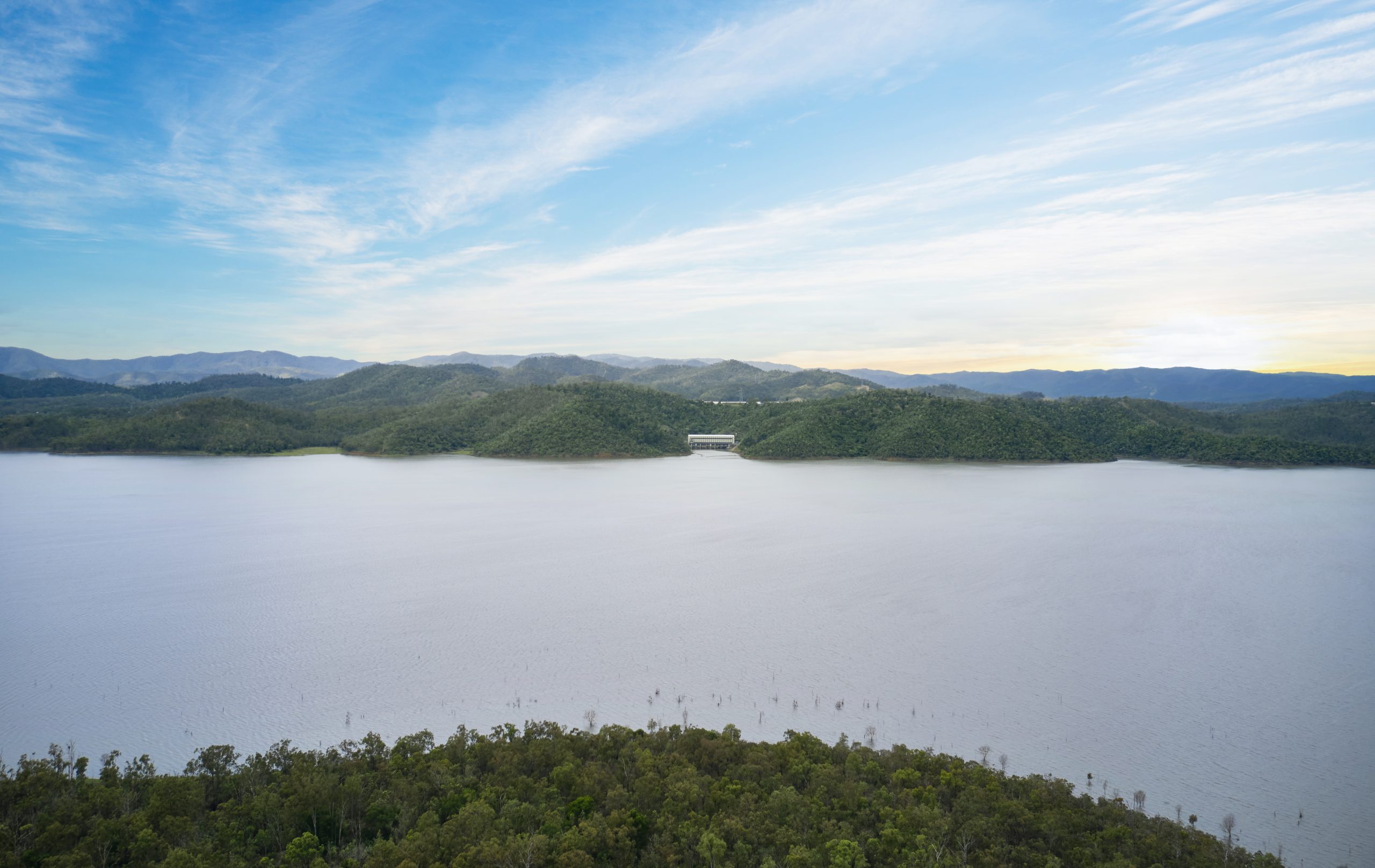 Large panoramic aerial view of Wivenhoe Dam showing the power station in the distance. It is beautiful, as the sun rises from behind the mountainous green landscape.