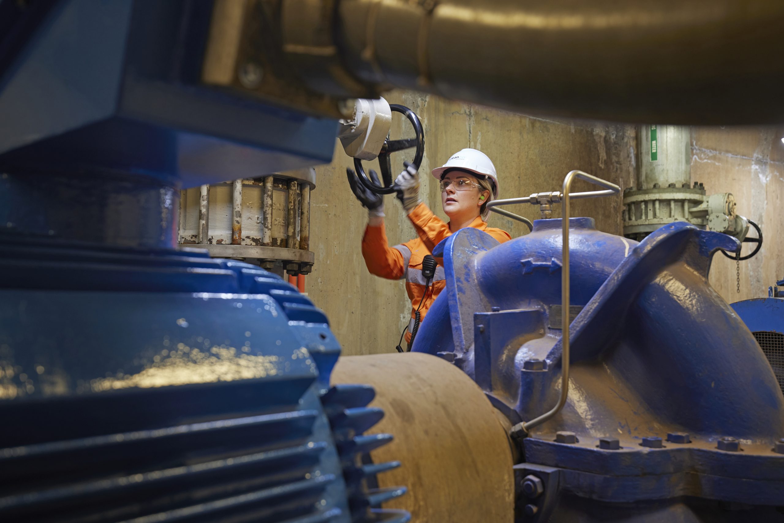 A close-up shot of a female CleanCo worker at the Wivenhoe Power Station. He is in between large blue pipes and turning on a pump.