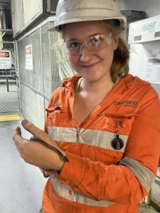 A CleanCo apprentice wearing high visibility clothing and a hard hat is standing, holding a small snake on her hand and smiling at the camera.