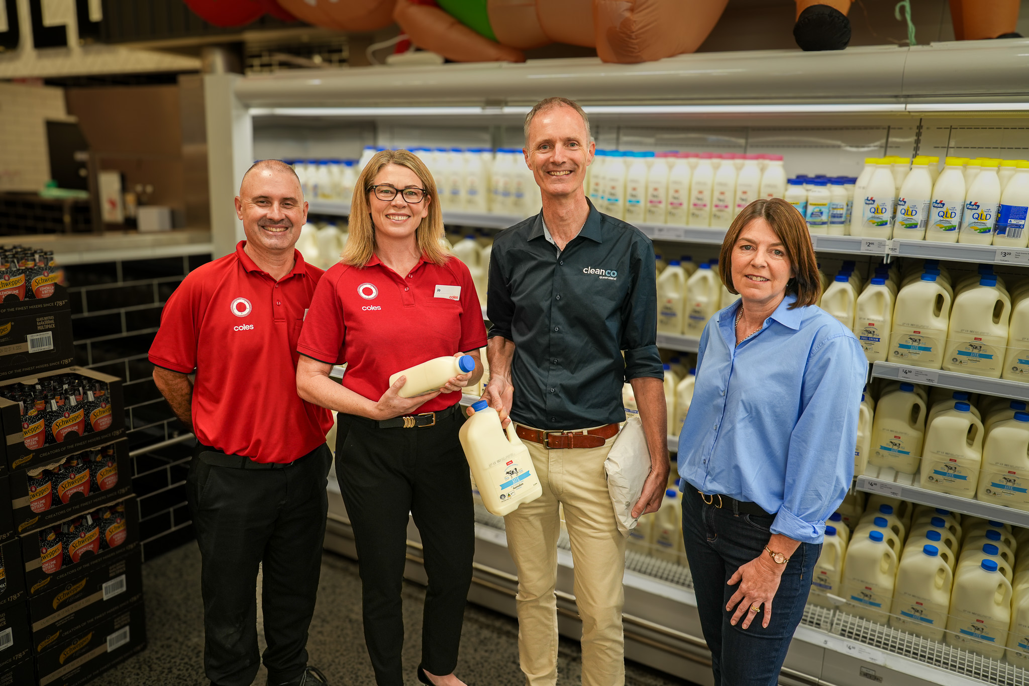 Two Coles employees stand with the CleanCo CEO and Board Chair in front of the cold section of a Coles supermarket, smiling at the camera. The CleanCo CEO is holding a bottle of milk.