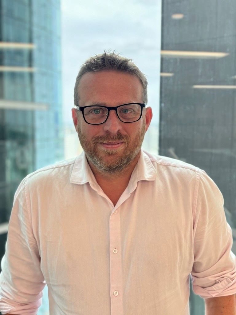 Trevor stands in front of a glass window, posing for his profile photo. He is wearing a white button up shirt and glasses.