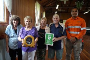 A CleanCo employee in high vis orange clothing stands with members of the Glamorgan Vale Community Hall Association. They are holding a defibrillator that CleanCo has donated to the hall.