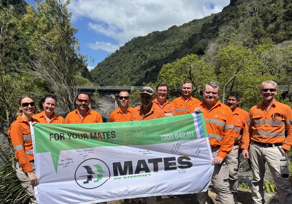 A group of CleanCo employees from Barron Gorge Power Station hold a Mates in Energy flag. They are standing with the Barron River behind them.