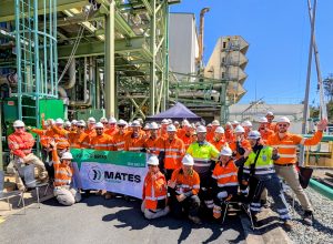 A large group of CleanCo employees and contractors stand in front of Swanbank E gas fired power station, cheering - they are holding a Mates in Energy flag